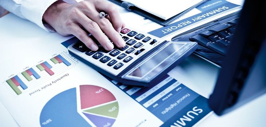 Photo of hand holding pen and using calculator at desk with computer and financial documents